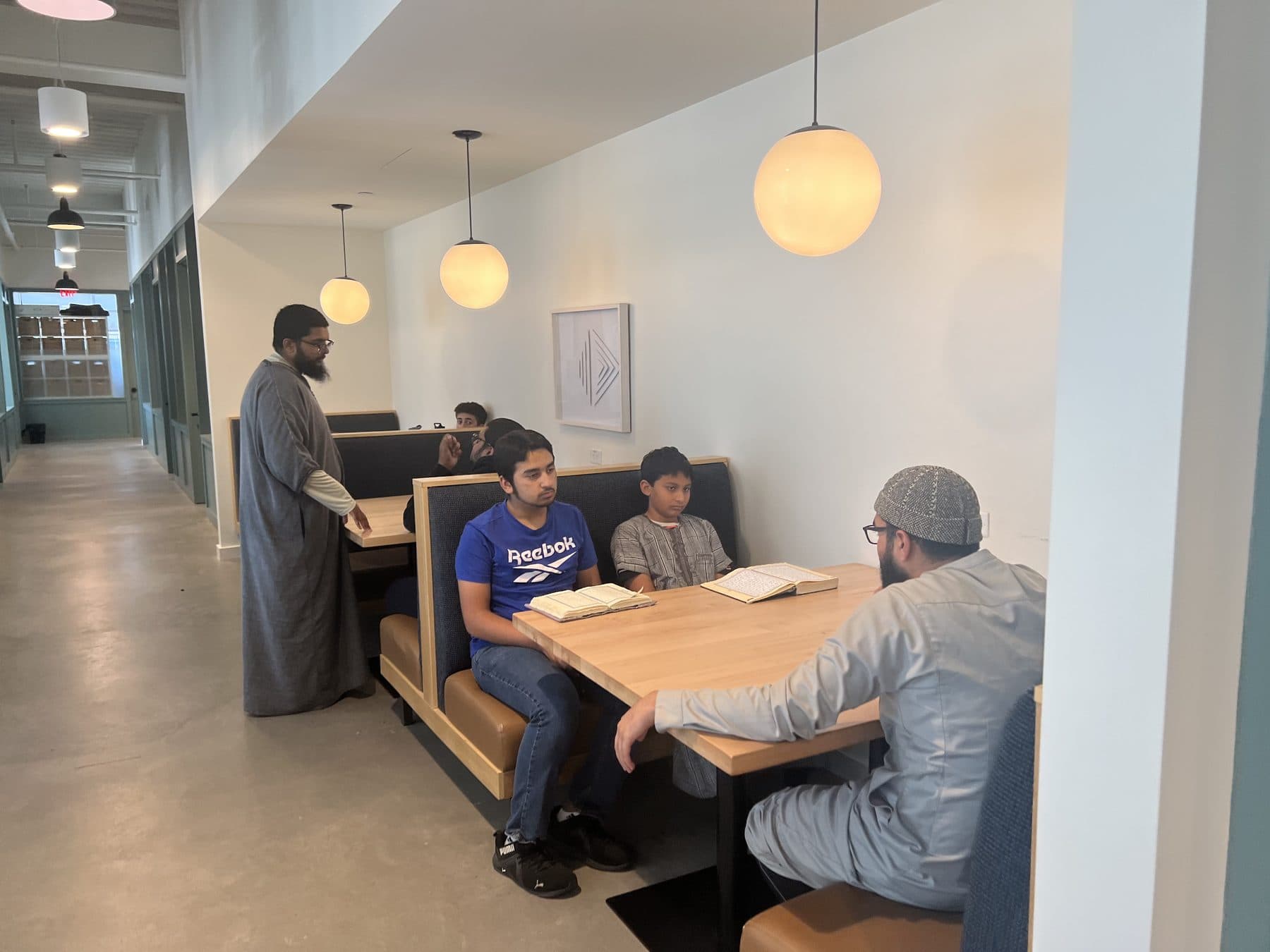 A teacher in a thobe speaking with two students at a table, a book open between them