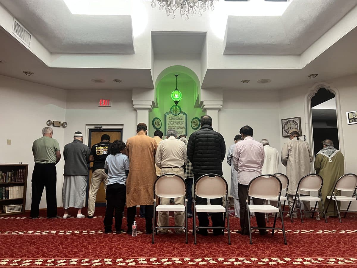 A congregation standing in night prayer beneath a green-lit mihrab