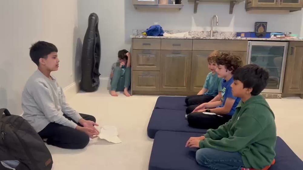 An older student sitting on the floor facing three younger boys seated on cushions during a circle