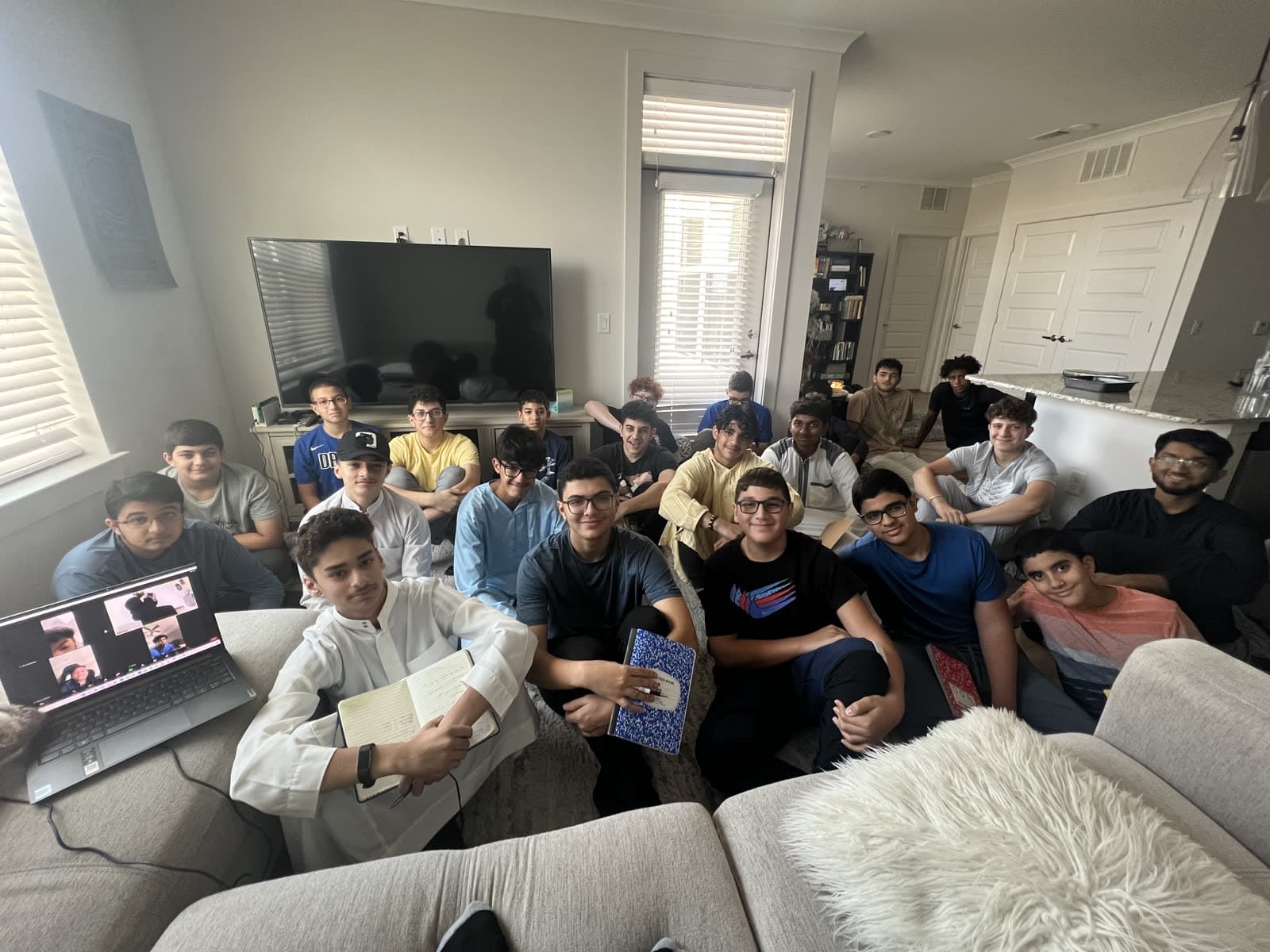 Around eighteen teenage boys seated together with notebooks during a weekly circle, a laptop beside them joining students online