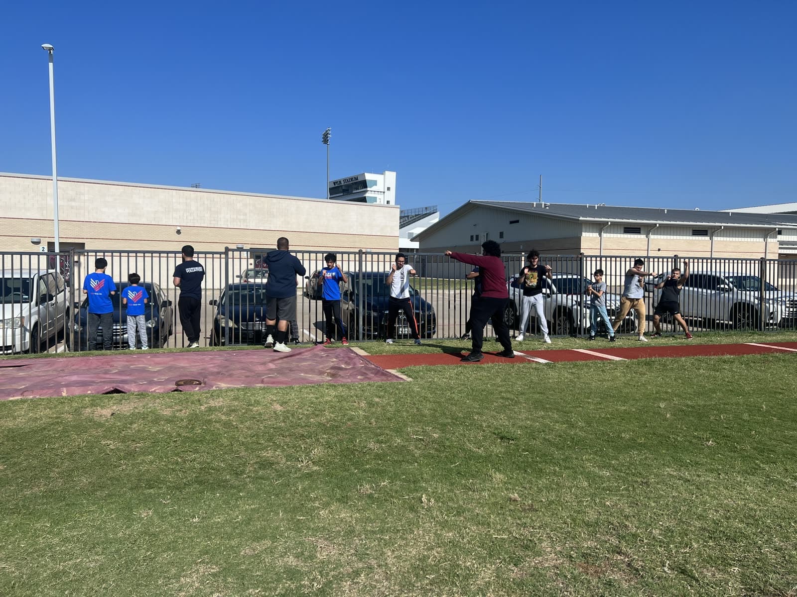 Boys lined up along the fence of a track field on a bright day out