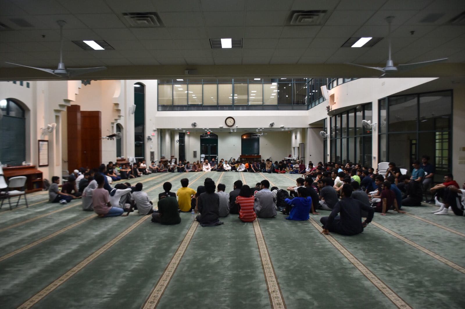 A wide circle of youth seated on the carpet of a large masjid prayer hall