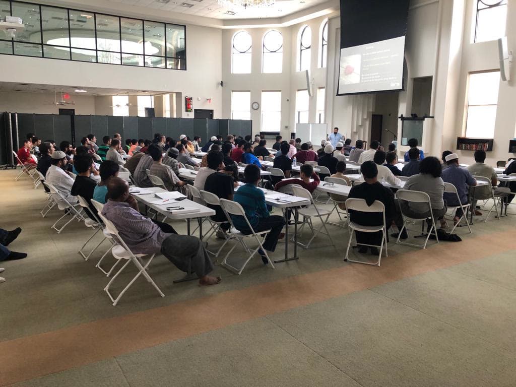 A large masjid hall filled with attendees seated for a program