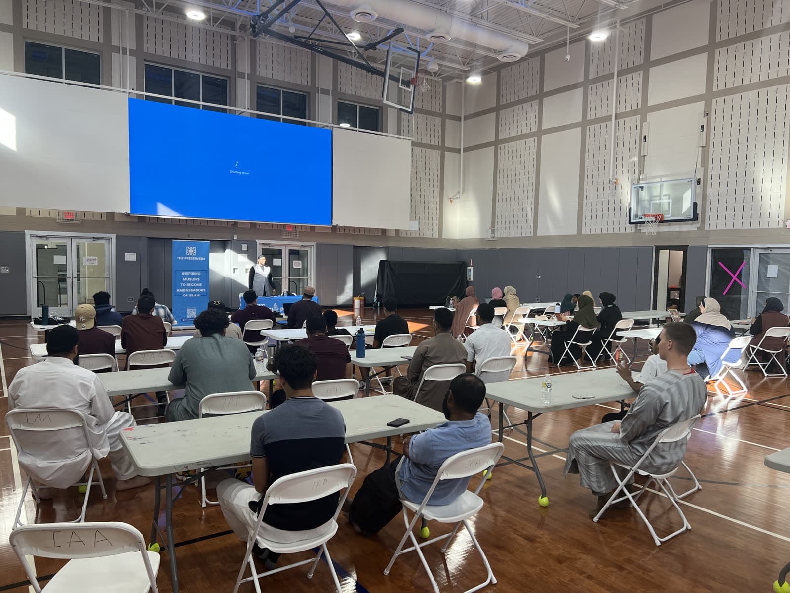 Young people seated at tables in a community hall during a workshop, a large screen behind the speaker
